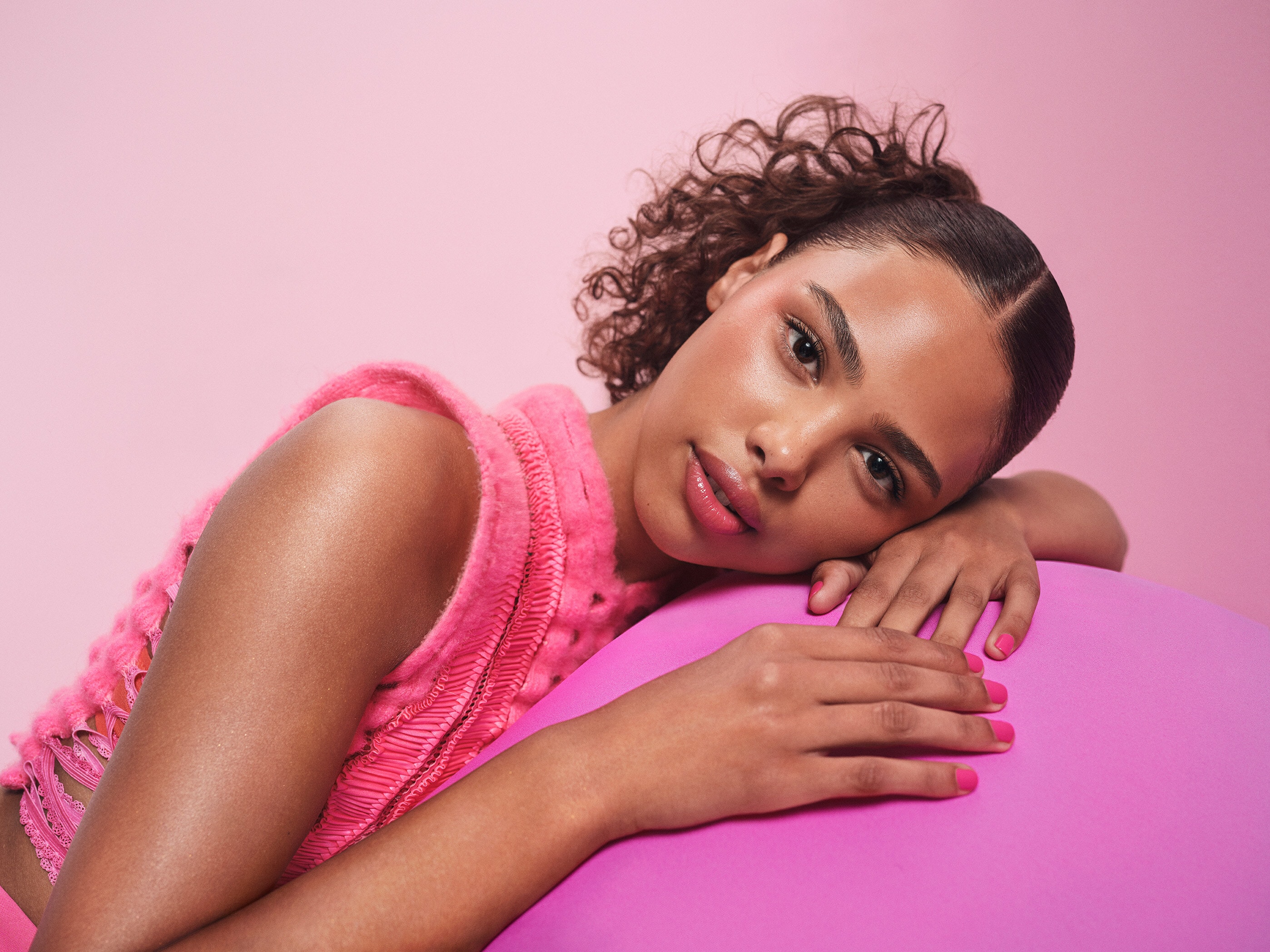 一名女性臉部靠在粉紅色圓形物品上,眼神柔和,臉上帶著粉紅色的 MAC 超漾光幻色嫩唇凍及超漾光持色奶凍腮紅,整體妝容顯得自然迷人。